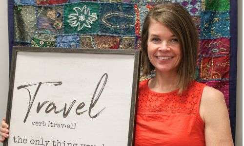 woman in red shirt standing in front of a colorful wall hanging and holding a sign