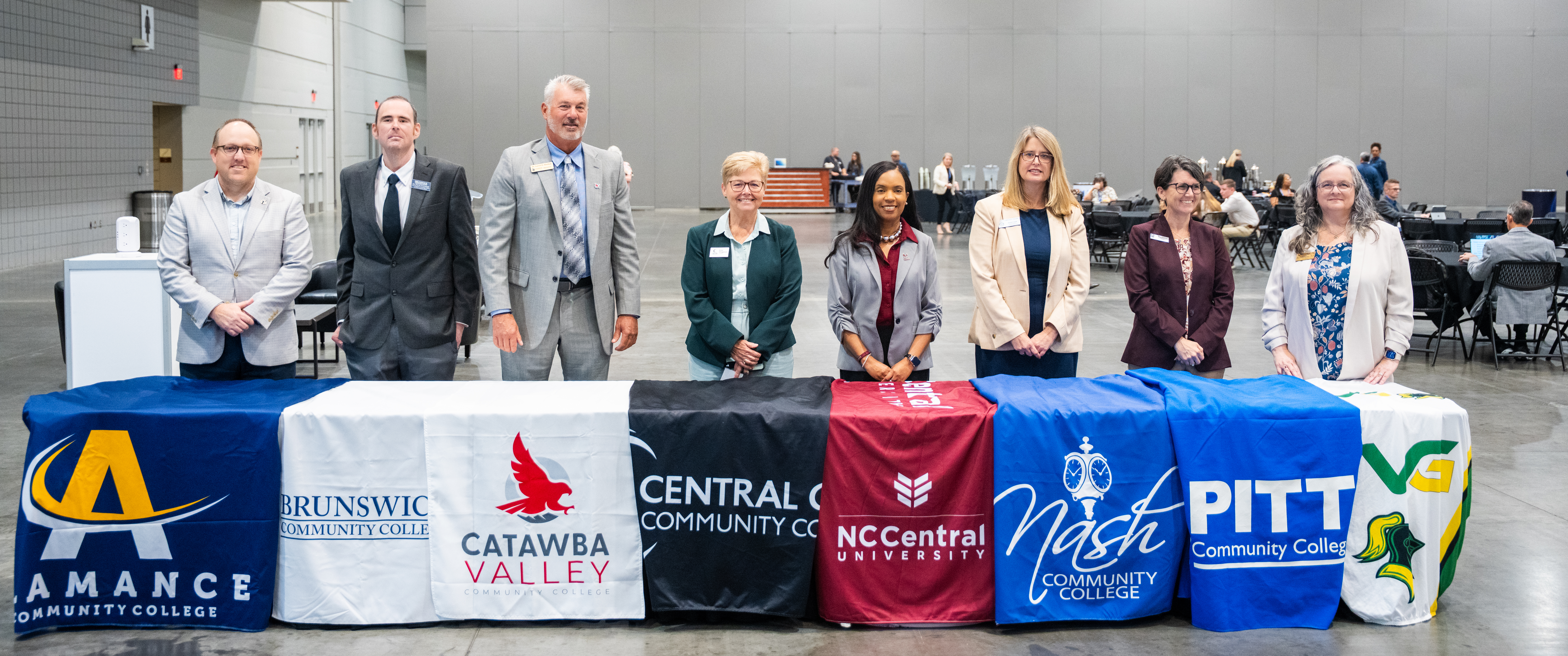 A group of eight individuals stands behind a row of tables covered with banners representing various community colleges.